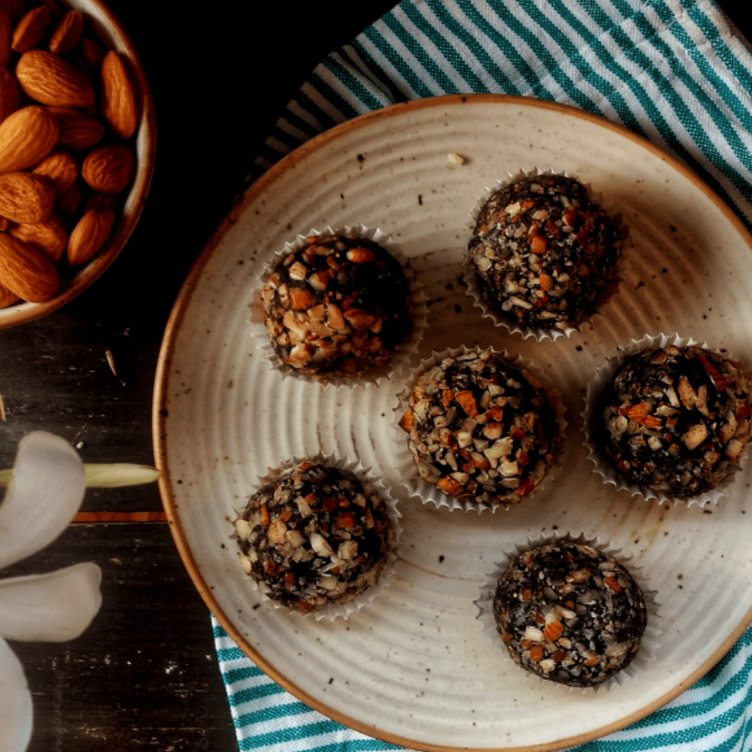 A plate with 6 Almond Fat Bombs by Fourmet, placed on a striped kitchen towel. A bowl full of almonds is partially visible in the top left corner.